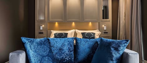Un dormitorio con un sofá gris, cojines estampados en azul, una cama con almohadas blancas y negras, y un espejo grande sobre la cama.