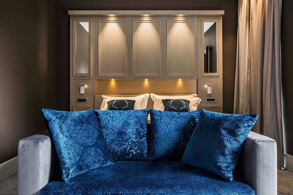 Un dormitorio con un sofá gris, cojines estampados en azul, una cama con almohadas blancas y negras, y un espejo grande sobre la cama.