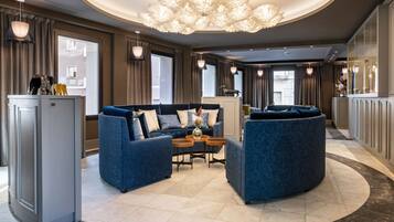 A spacious lobby with a central seating area featuring blue sofas and a round table, a modern chandelier, and a view of the outside through large windows.