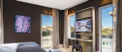 A modern hotel room with a bed, a desk, a TV, and a balcony with a view of buildings.