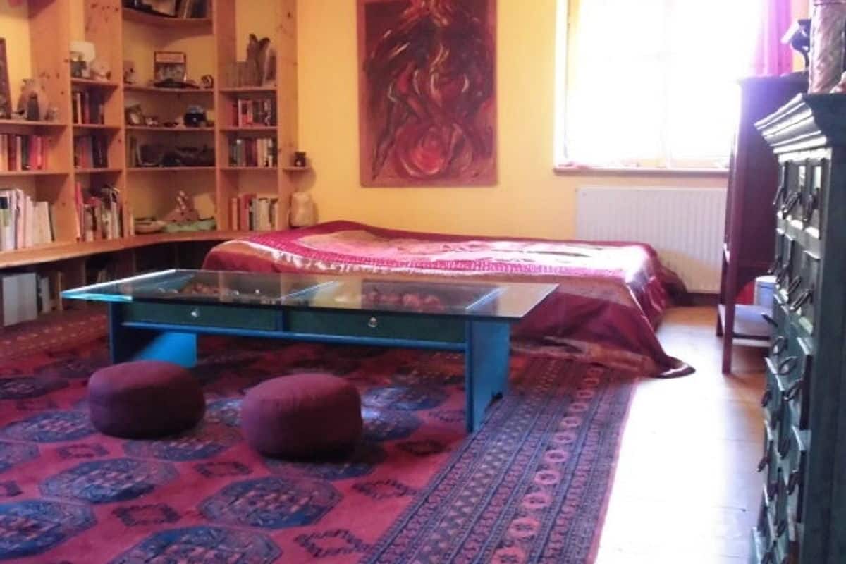 Une pièce avec une table à plateau en verre, deux poufs rouges, une couvre-lit rouge et une bibliothèque remplie de livres.