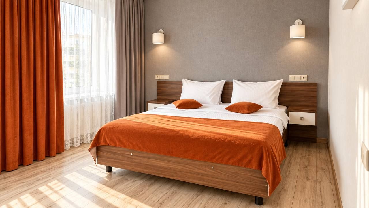 Un lit double avec un cadre en bois et une literie orange, deux tables de chevet avec des lampes, une fenêtre avec des rideaux et un mur de couleur claire.