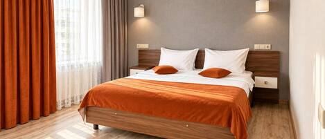 A double bed with a wooden frame and orange bedding, two bedside tables with lamps, a window with curtains, and a light-colored wall.
