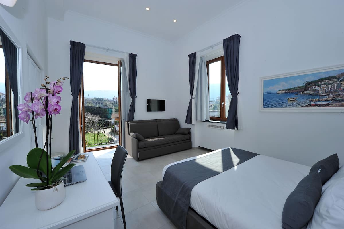 A modern hotel room with a bed, a desk with a laptop, a sofa, and a view of the sea through large windows.