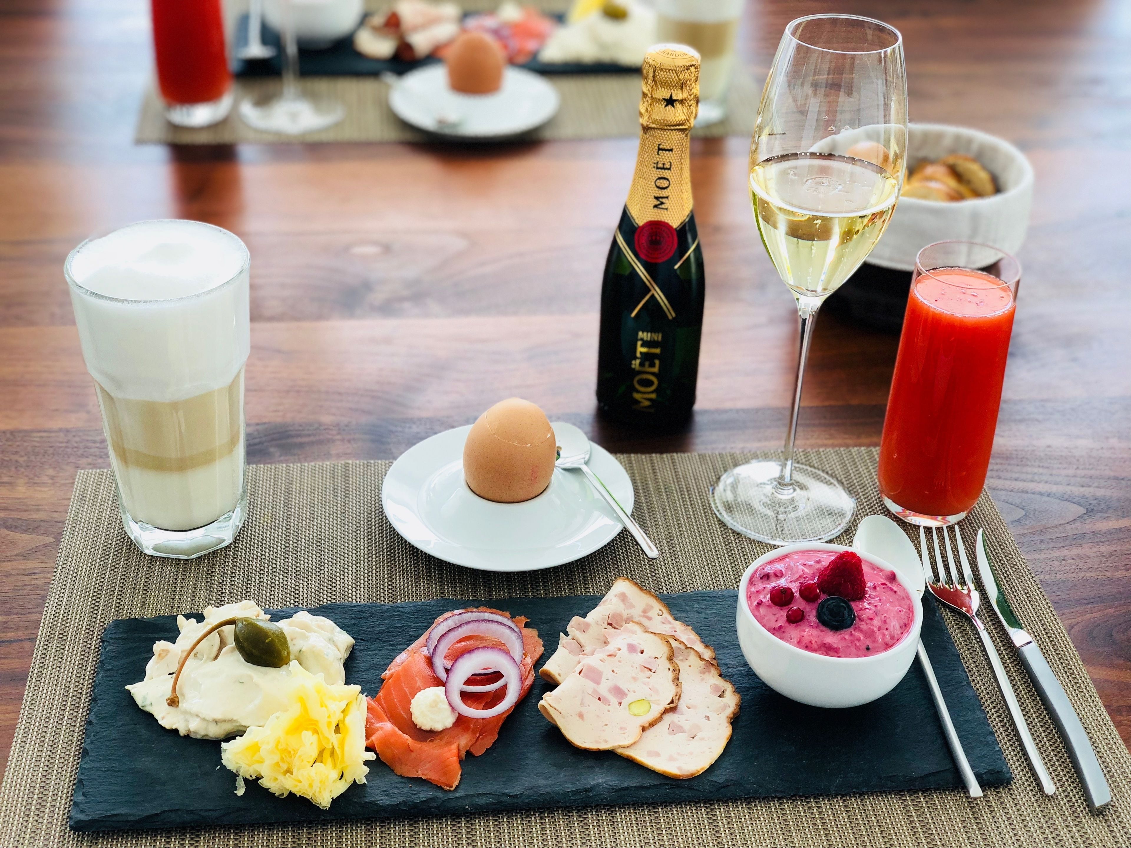 A dining table with a variety of food items, including smoked salmon, cheese, and a bowl of fruit salad, along with a bottle of champagne and glasses of wine.