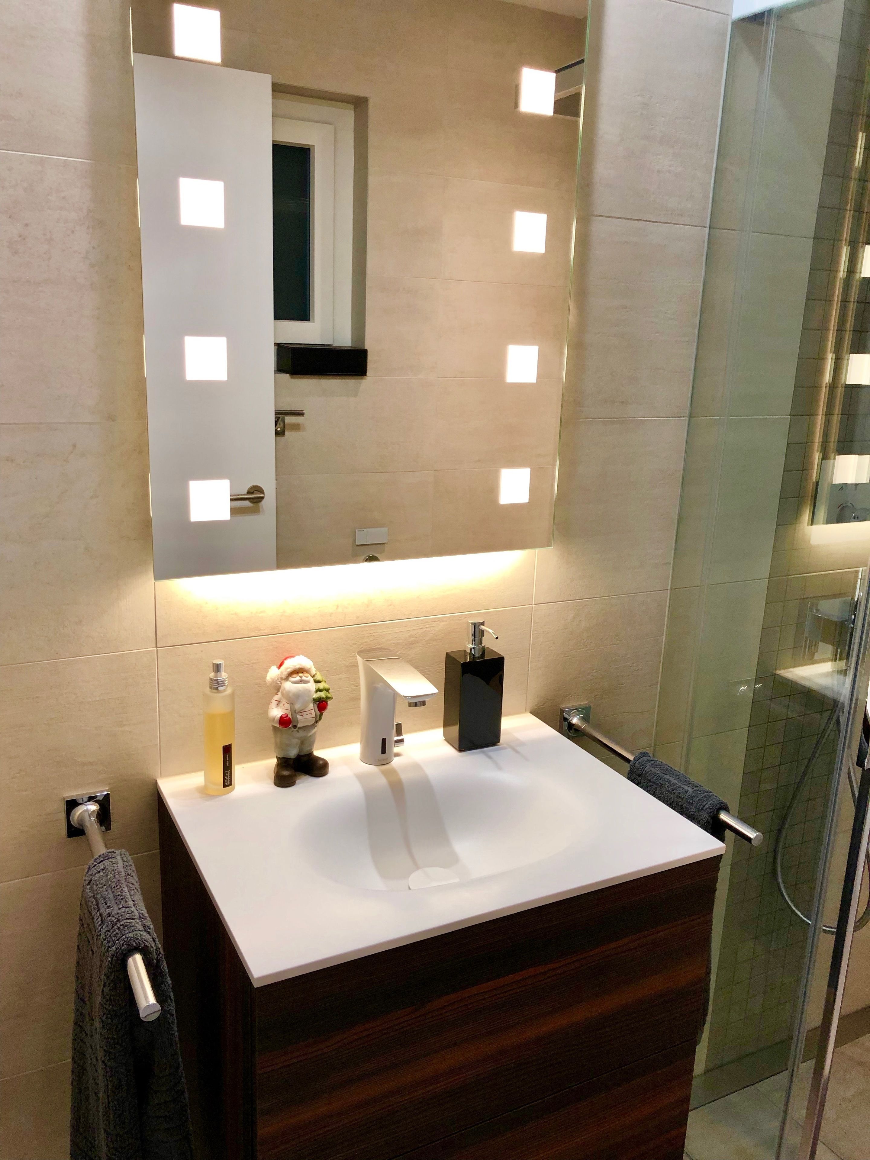 A bathroom with a white vanity, a mirror with square lights, a towel rack, a bottle of liquid soap, and a small Santa figurine.