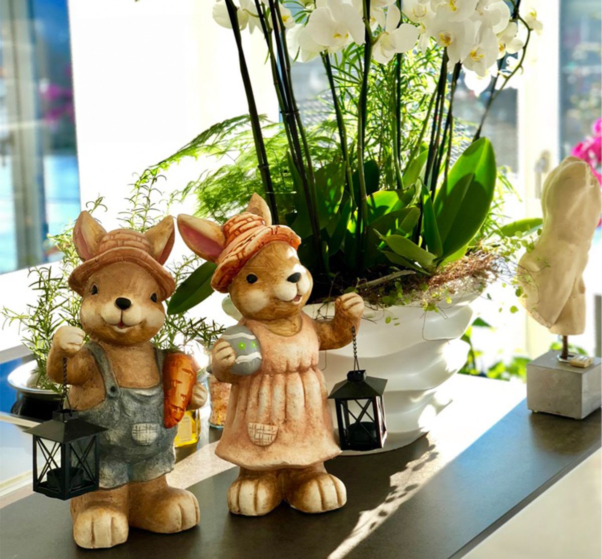 Twee keramieken konijnenbeeldjes in een witte pot met groene planten en witte bloemen.
