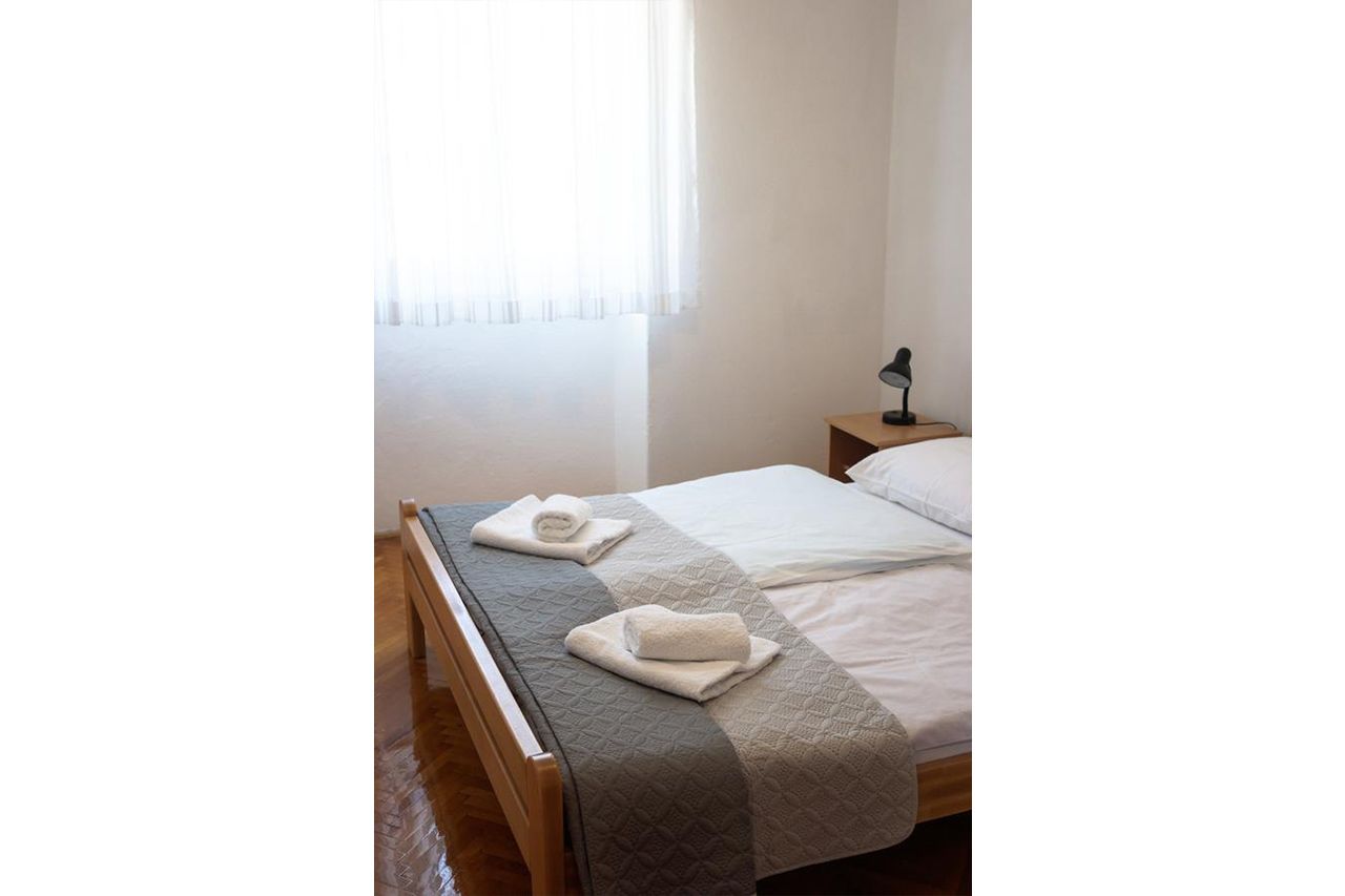 Uma cama bem arrumada com um cobertor acolchoado e duas toalhas enroladas, um abajur e uma janela com cortinas leves.