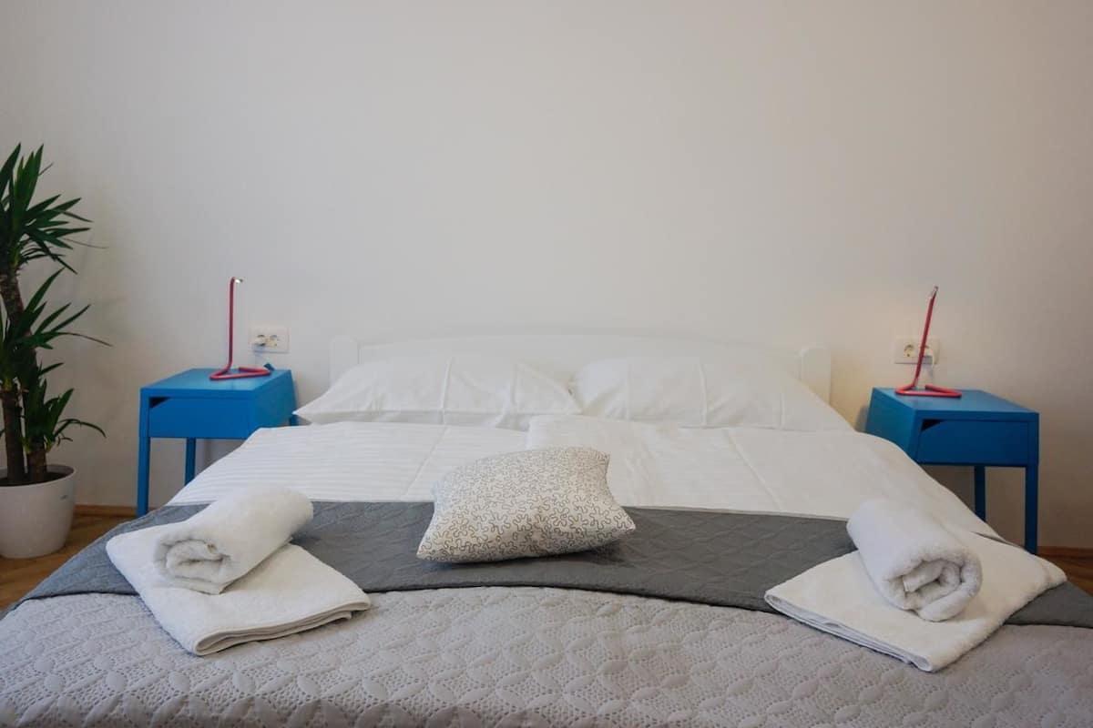 Un lit bien fait, avec du linge de lit blanc, une couverture grise et deux tables de chevet bleues, sur lesquelles sont posées des lampes rouges.
