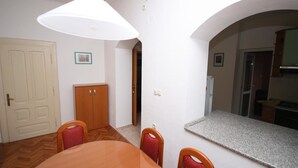A dining area with a wooden table and chairs, a white ceiling lamp, and a kitchen visible through an archway.