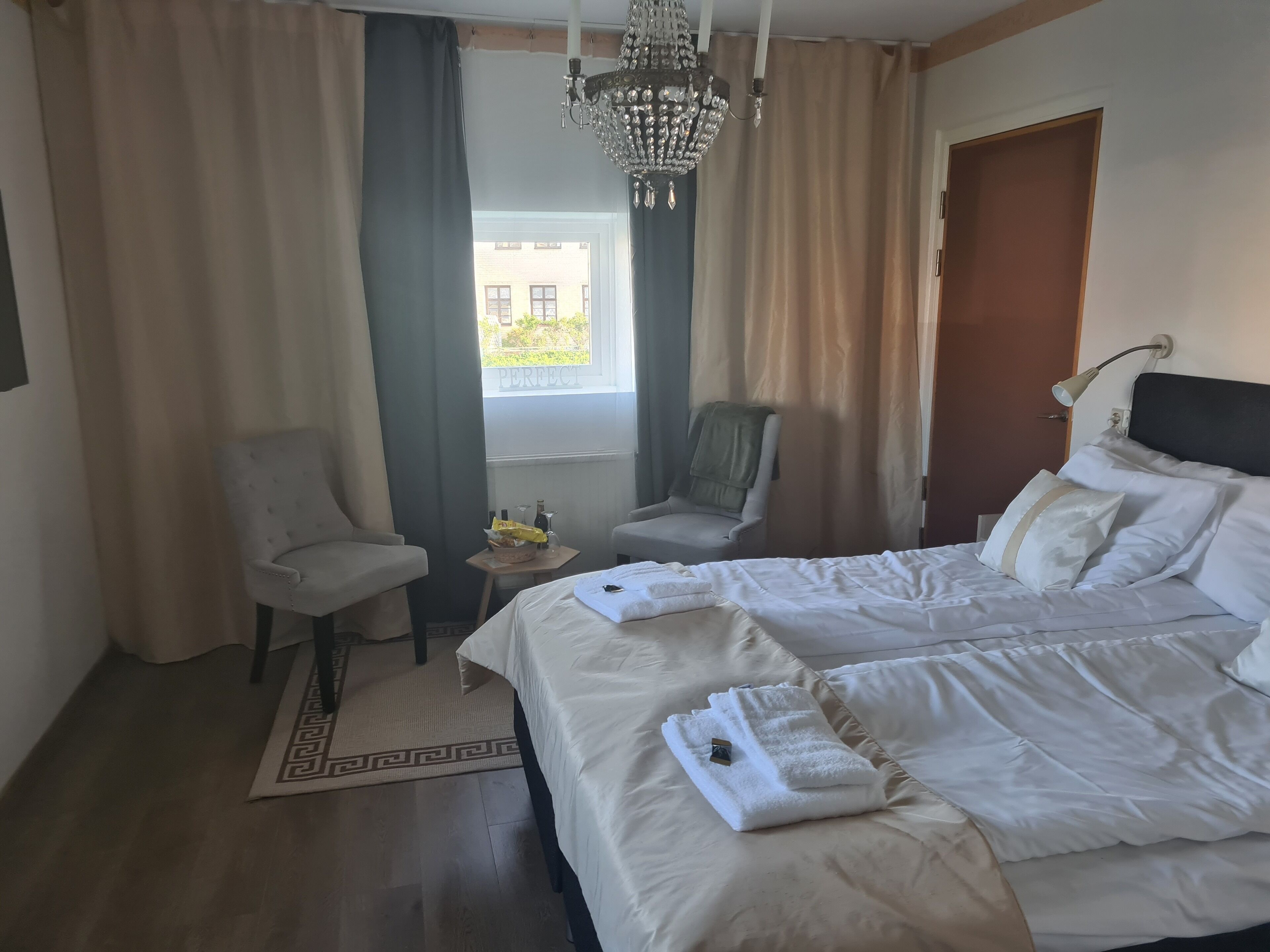 A hotel room with a large bed, two armchairs, a small table with a bottle and a glass, and a chandelier.