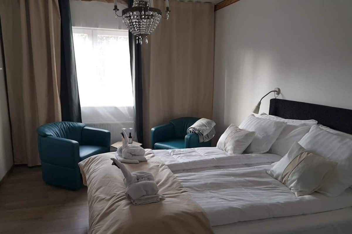 Quarto de hotel com uma cama grande, duas poltronas azuis, um candelabro e vista para o exterior através de uma janela com cortinas.