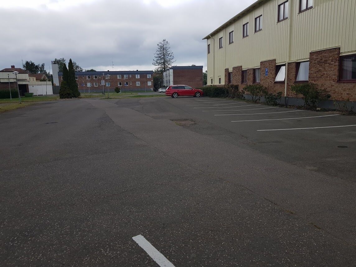 A parking lot with a red car parked in the distance, surrounded by buildings with brick and metal exteriors.
