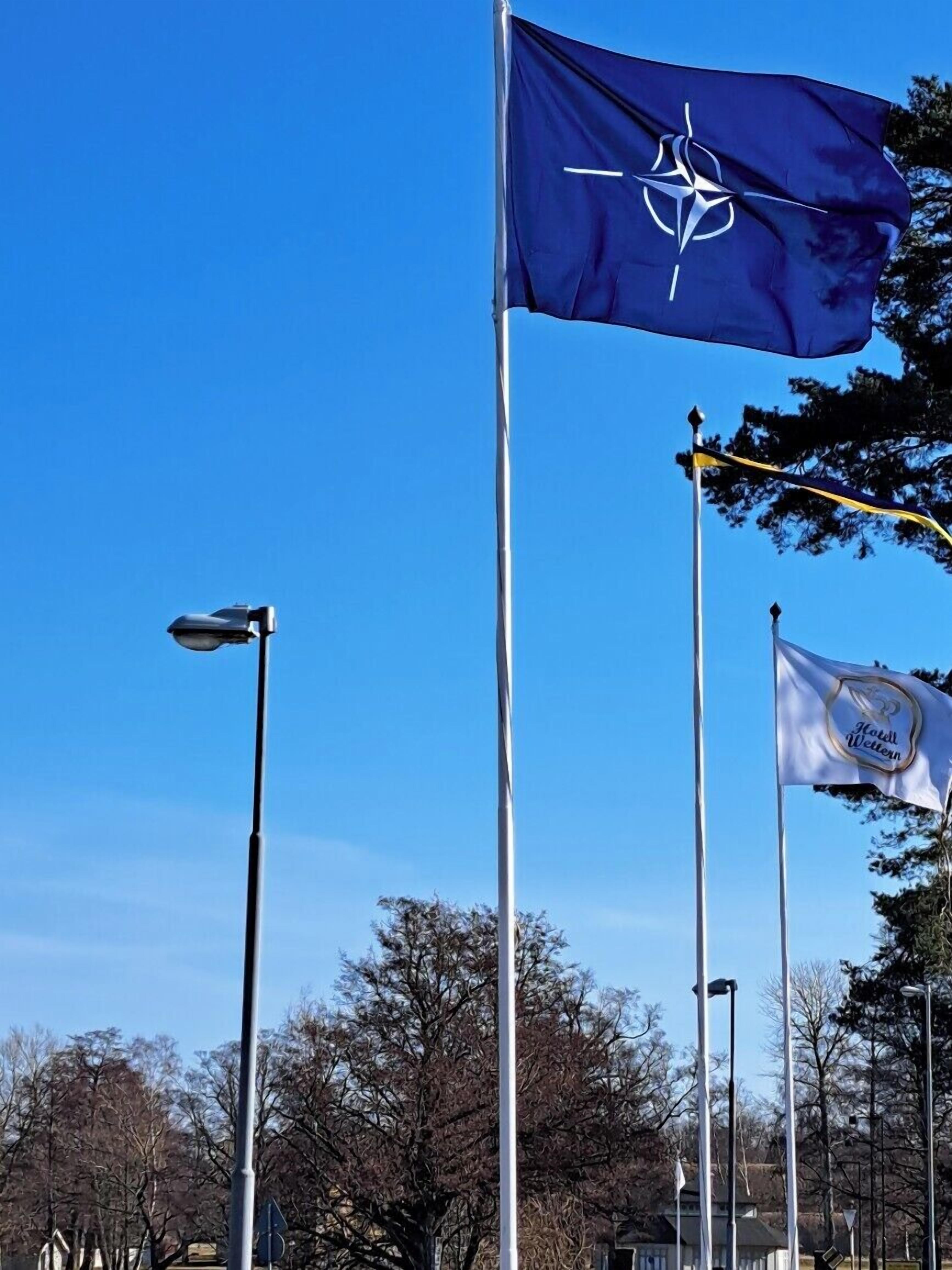 Eine Fahnenmasten mit einer blauen Flagge mit einem weißen Kompassmuster, einer weißen Flagge mit einem Logo und einer Straßenlaterne.
