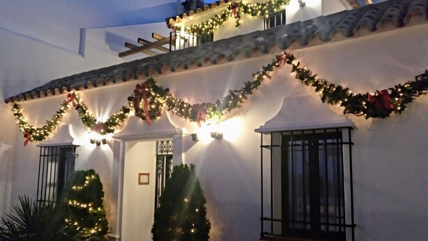 Un edificio con fachada blanca, decorado con luces navideñas y guirnaldas. Se observan dos ventanas con rejas de hierro negro y una puerta con una pequeña placa en su parte superior.