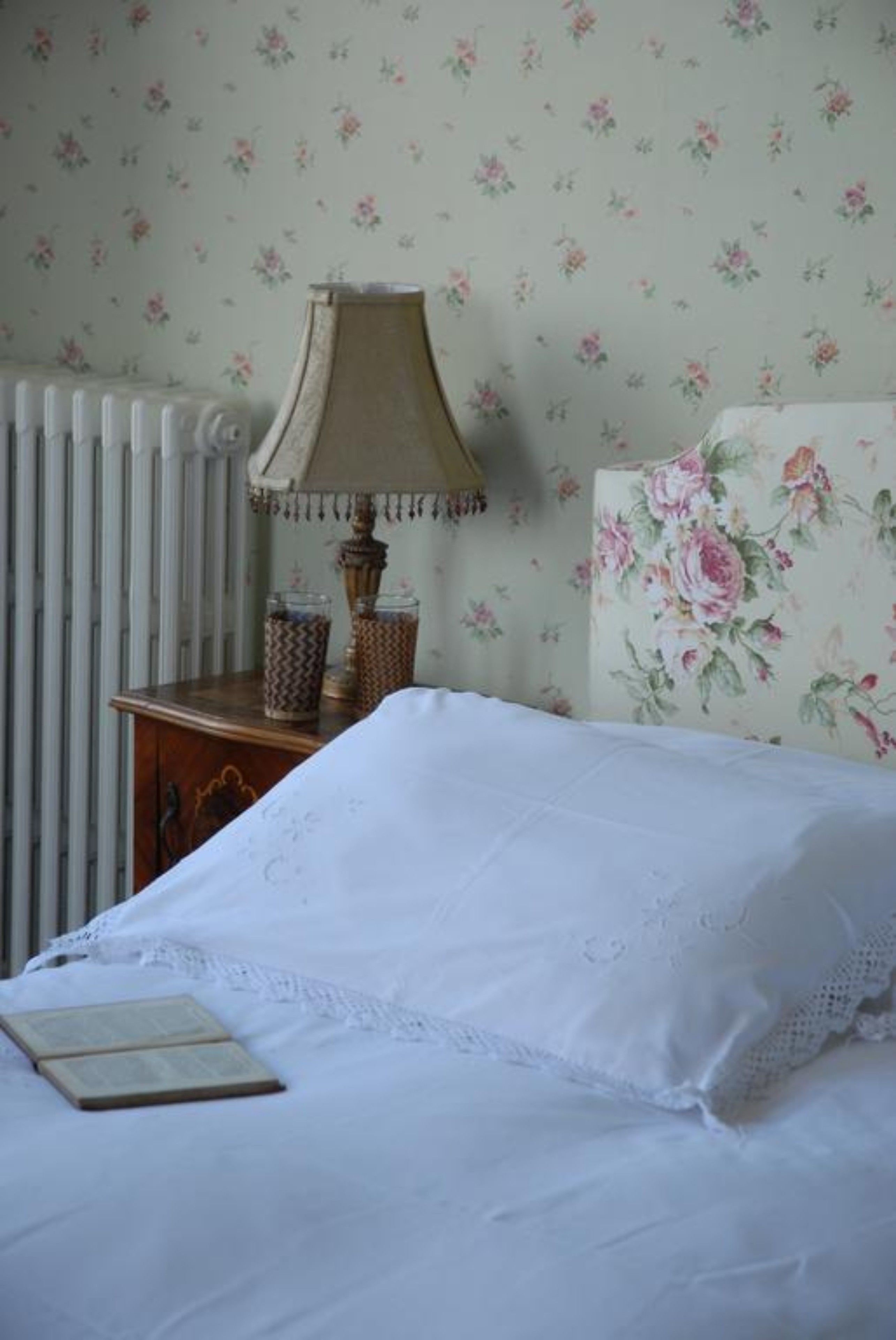 A bedroom with a bed, a nightstand with a lamp, a radiator, and floral wallpaper.