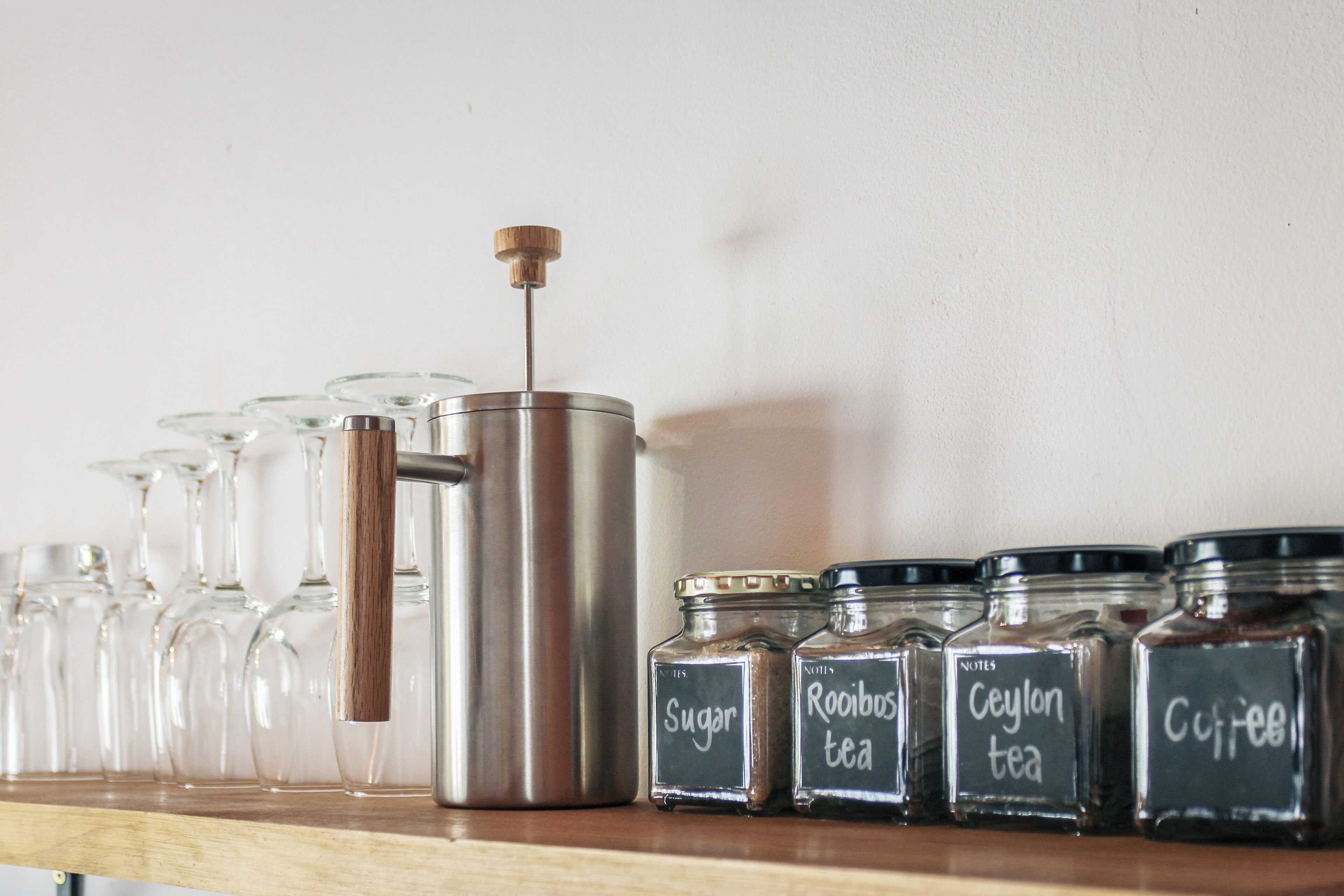 A kitchen shelf with a coffee press, sugar, rooibos tea, and ceylon tea containers.
