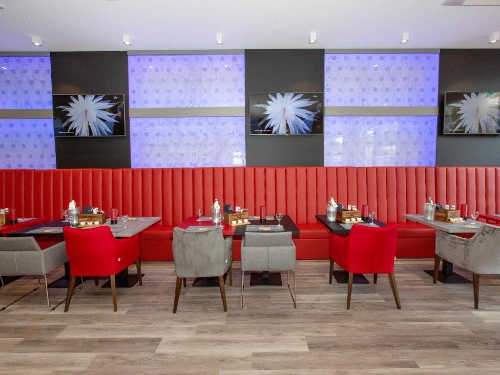 Ein Restaurant mit rot gepolsterten Sitzecken und grauen Stühlen, das ein modernes Design mit gemustertem Wandpapier und Deckenleuchten aufweist.