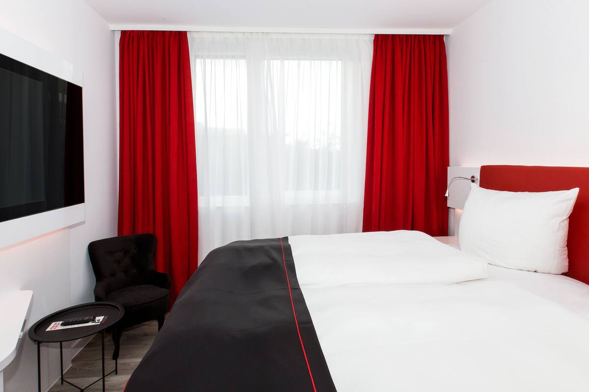 Habitación de hotel con una cama grande, televisor montado en la pared, escritorio con silla y cortinas rojas.