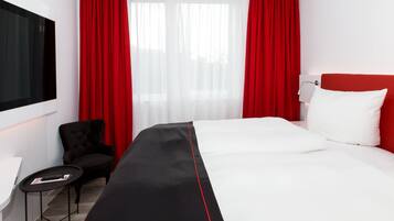 A hotel room with a large bed, a TV mounted on the wall, a desk with a chair, and red curtains.