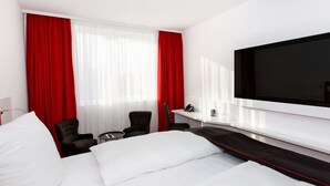 A hotel room with a large bed, a desk, two chairs, a TV, and red curtains.