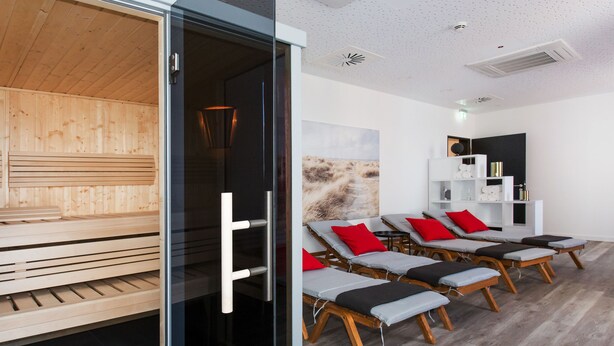 A modern spa room with wooden floors, glass doors, and multiple sun loungers with red cushions.