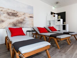 Un cuarto de spa con tres camillas de madera para masajes, cada una con un cojín gris y una almohada roja. Una gran pintura abstracta decora la pared detrás de las camillas.