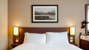 Una cama bien hecha con ropa blanca, dos lámparas de noche y una fotografía enmarcada de un paisaje en la pared.