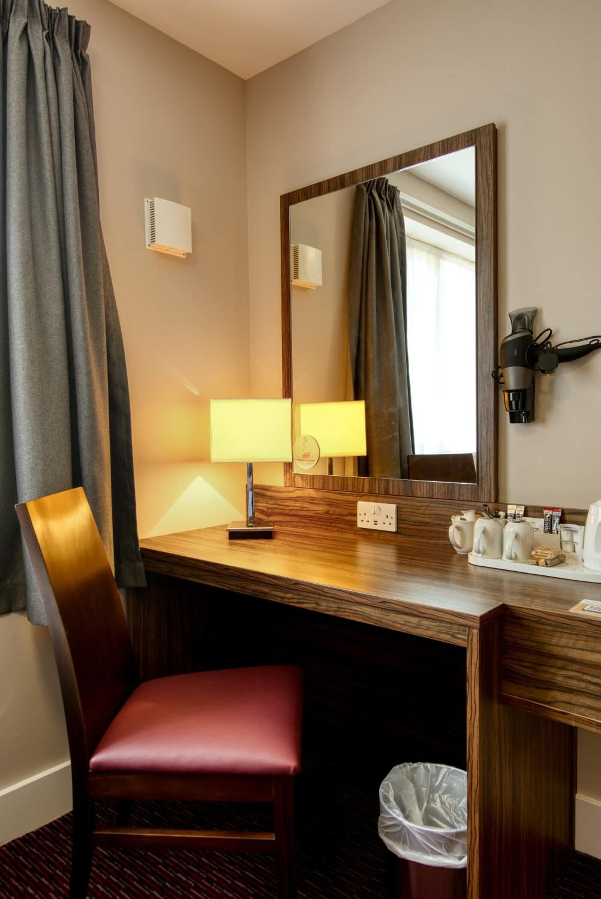 A hotel room with a wooden desk, a red chair, a mirror, a lamp, a hairdryer, and a trash bin.