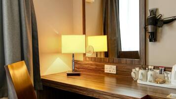 A hotel room with a wooden desk, a red chair, a mirror, a lamp, a hairdryer, and a trash bin.