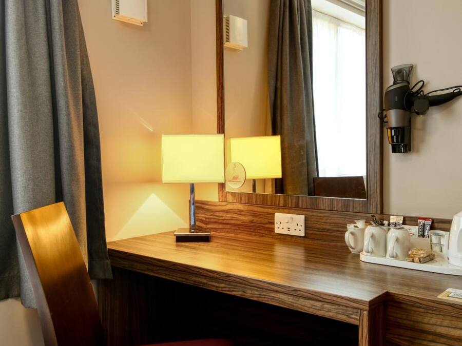 Ett hotellrum med ett skrivbord i trä, en röd stol, en spegel, en lampa, en hårtork och en papperskorg.