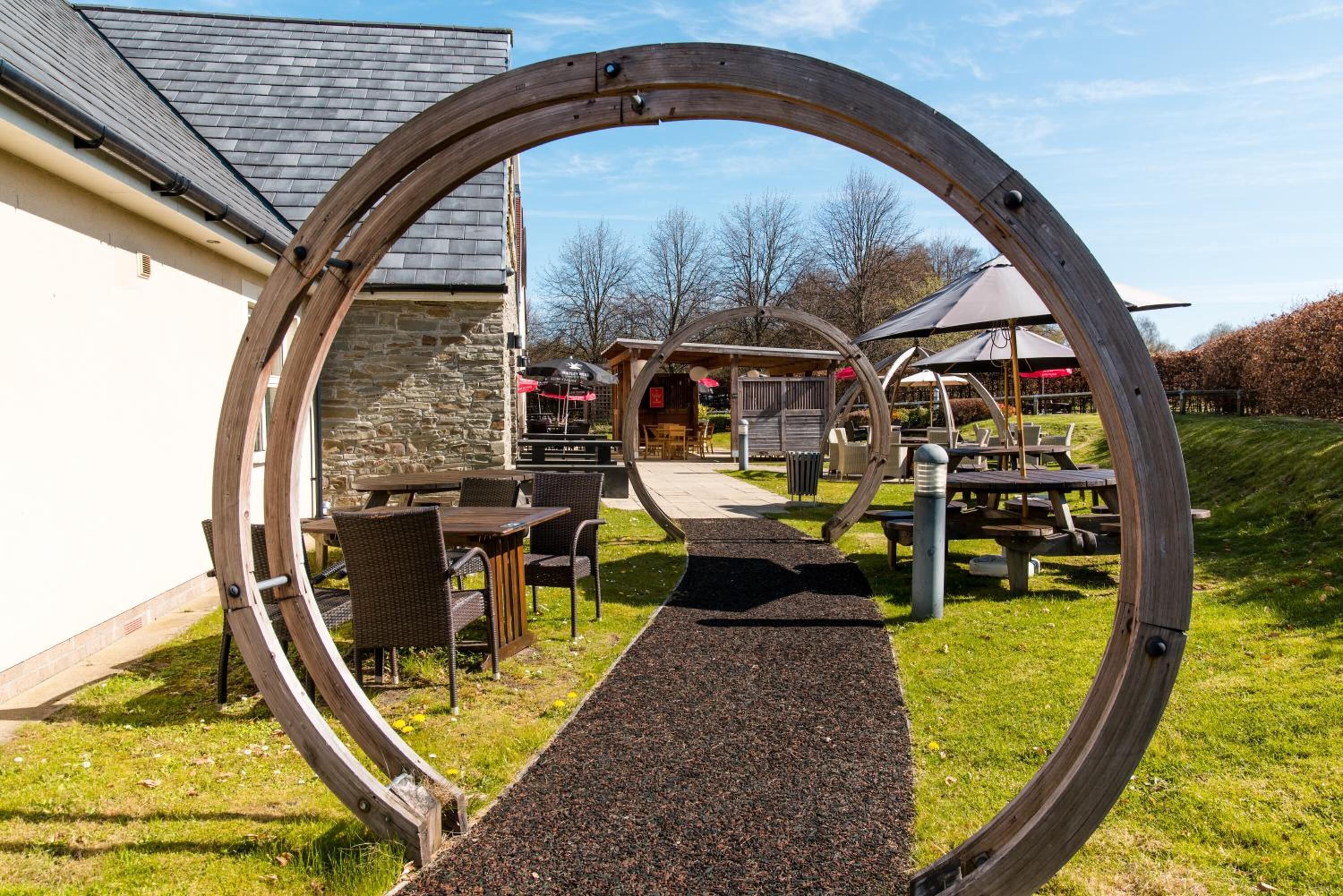 Une structure circulaire en bois encadre un chemin menant à un bâtiment doté d’une terrasse et d’un espace couvert.