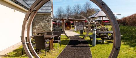 Une structure circulaire en bois encadre un chemin menant à un bâtiment doté d’une terrasse et d’un espace couvert.