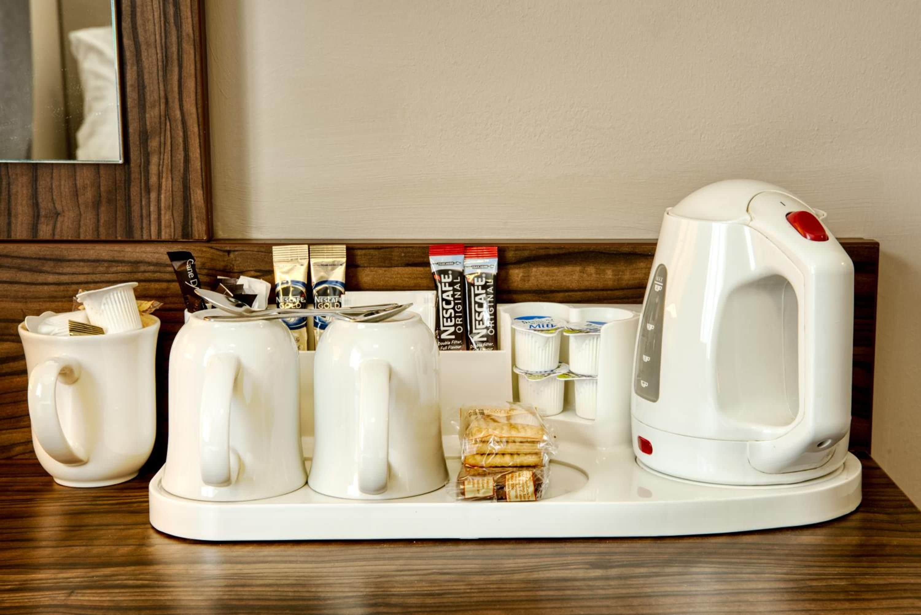 A hotel minibar setup with a coffee maker, two mugs, a glass, and various food and drink items.
