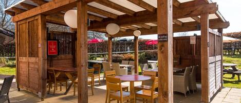 Un espace extérieur aménagé avec des tables et des chaises en bois, une zone couverte avec un panneau indicateur et un tableau affichant le menu.