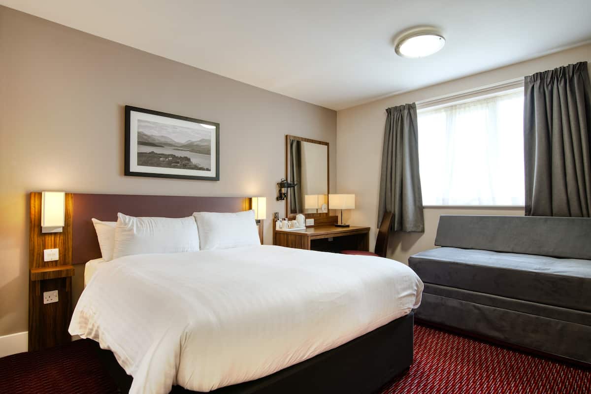 Habitación de hotel con una cama grande, un sofá, un escritorio con lámpara, un espejo y una ventana con cortinas.