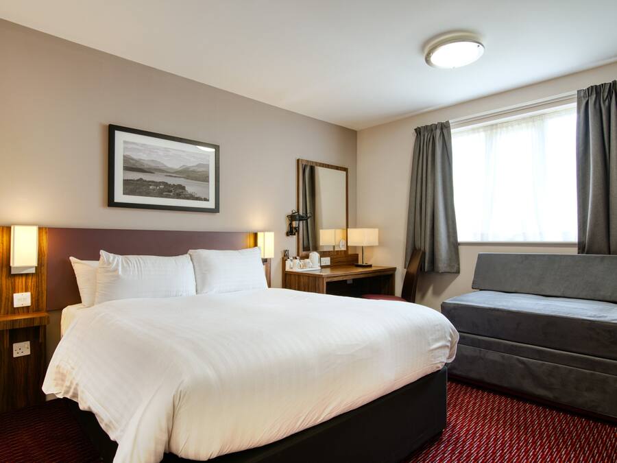 Habitación de hotel con una cama grande, un sofá, un escritorio con lámpara, un espejo y una ventana con cortinas.