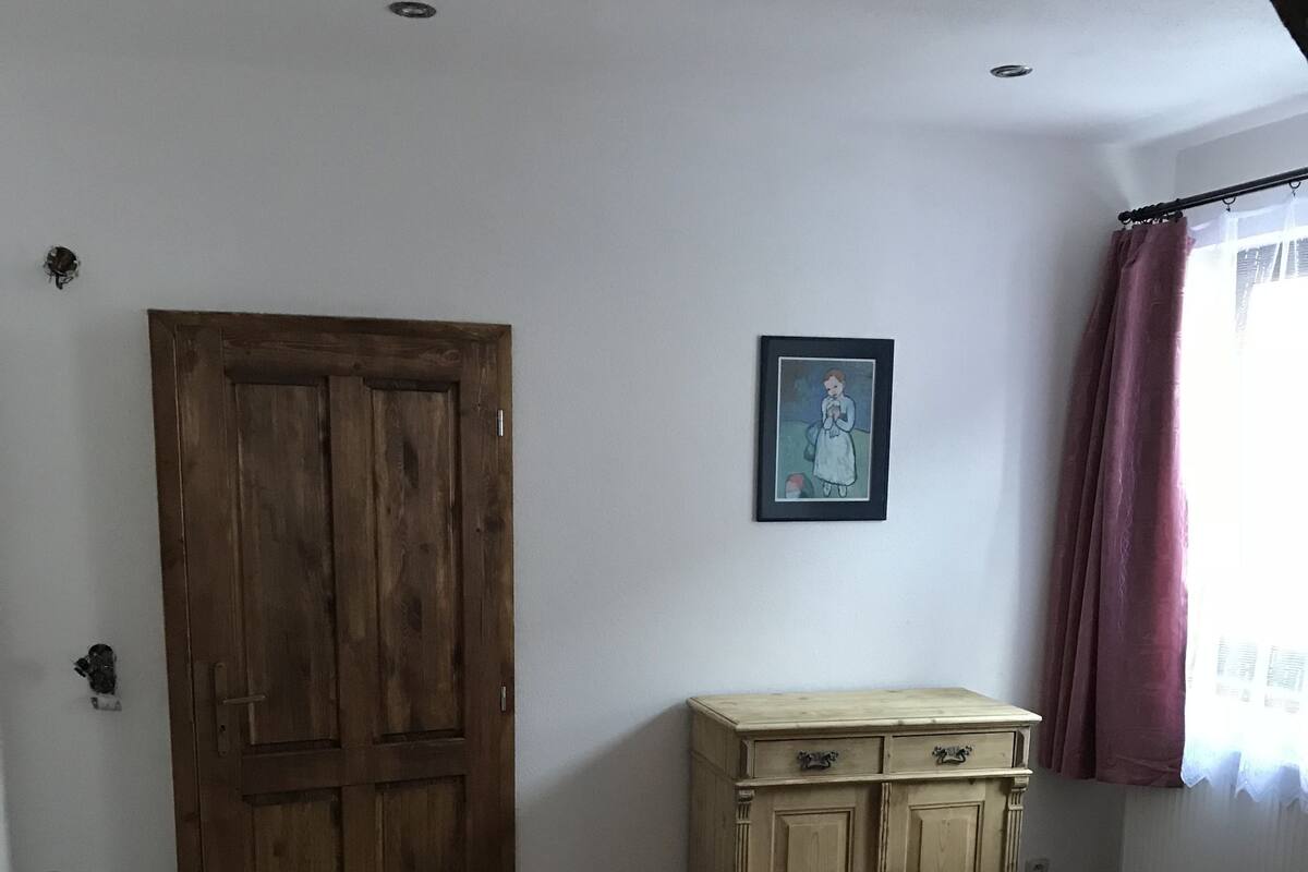 Un dormitorio con un armario de madera, una cama con edredón rosa estampado, una mesita de noche pequeña con una lámpara y un cuadro en la pared.