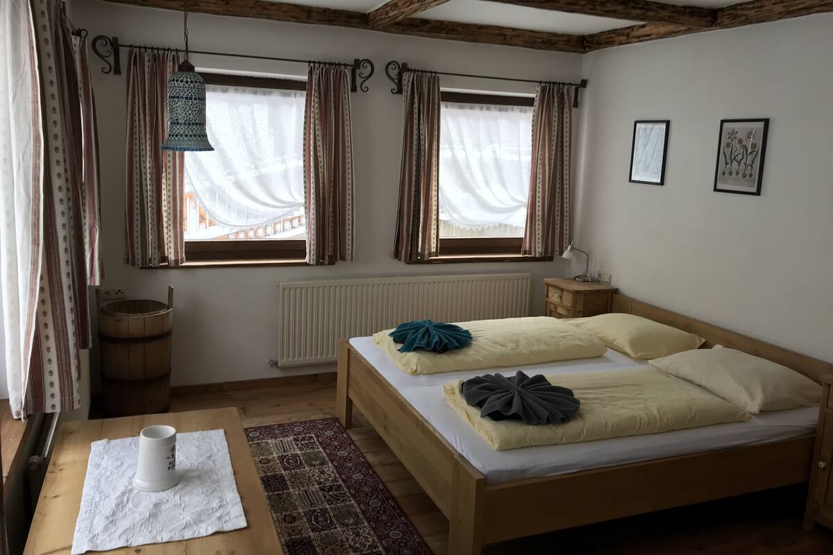 Un dormitorio acogedor con una cama de madera, dos ventanas con cortinas, un radiador, una mesita de noche de madera y una mesa con una taza blanca.