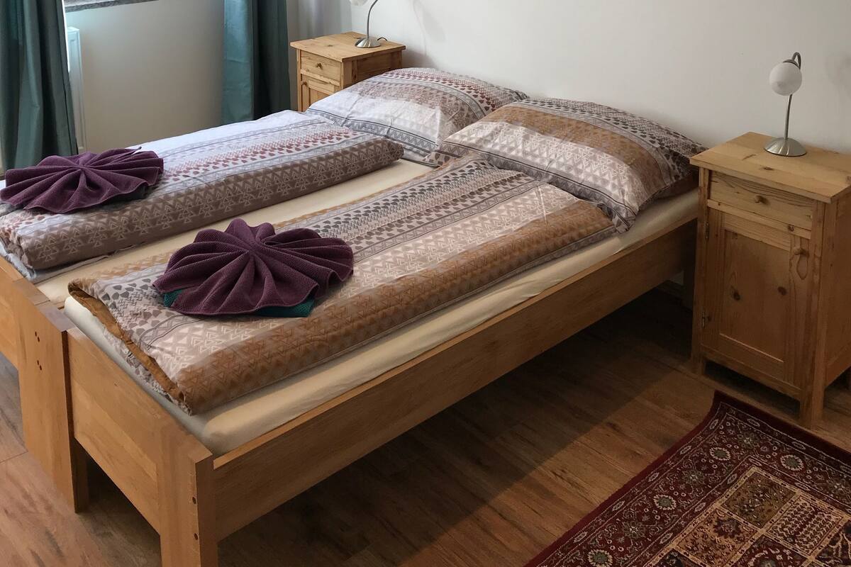 Una cama doble con ropa de cama estampada, una mesita de noche con lámpara, un pequeño armario de madera y un cuadro de un paisaje montañoso en la pared.