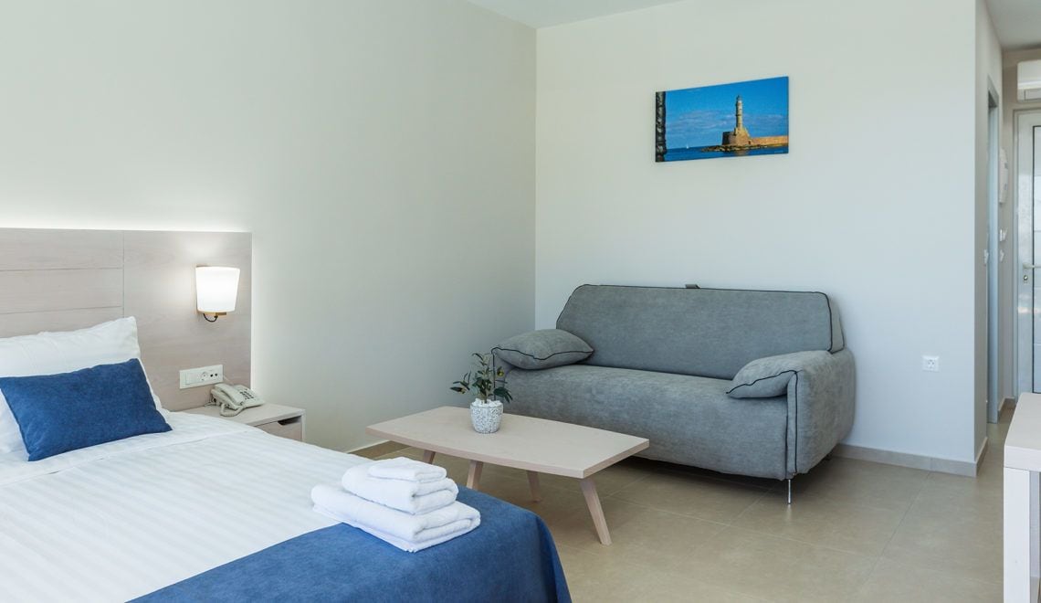 Habitación de hotel con una cama, un sofá gris, una mesita con un jarrón y un cuadro en la pared.
