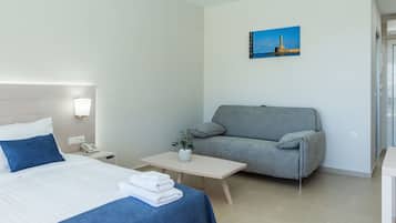 Habitación de hotel con una cama, un sofá gris, una mesita con un jarrón y un cuadro en la pared.