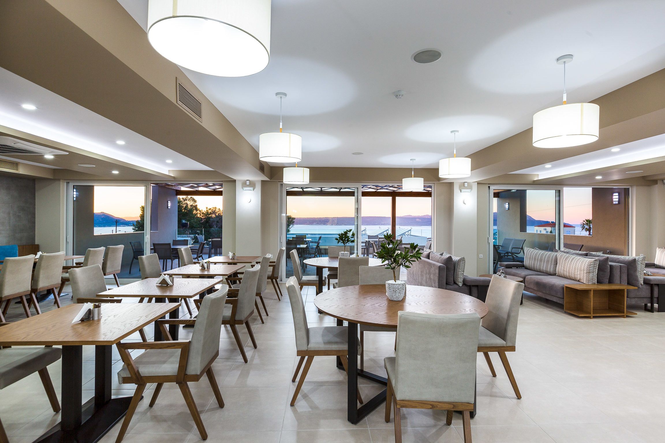 A modern dining area with tables and chairs, large windows offering a view of the sea, and pendant lighting.