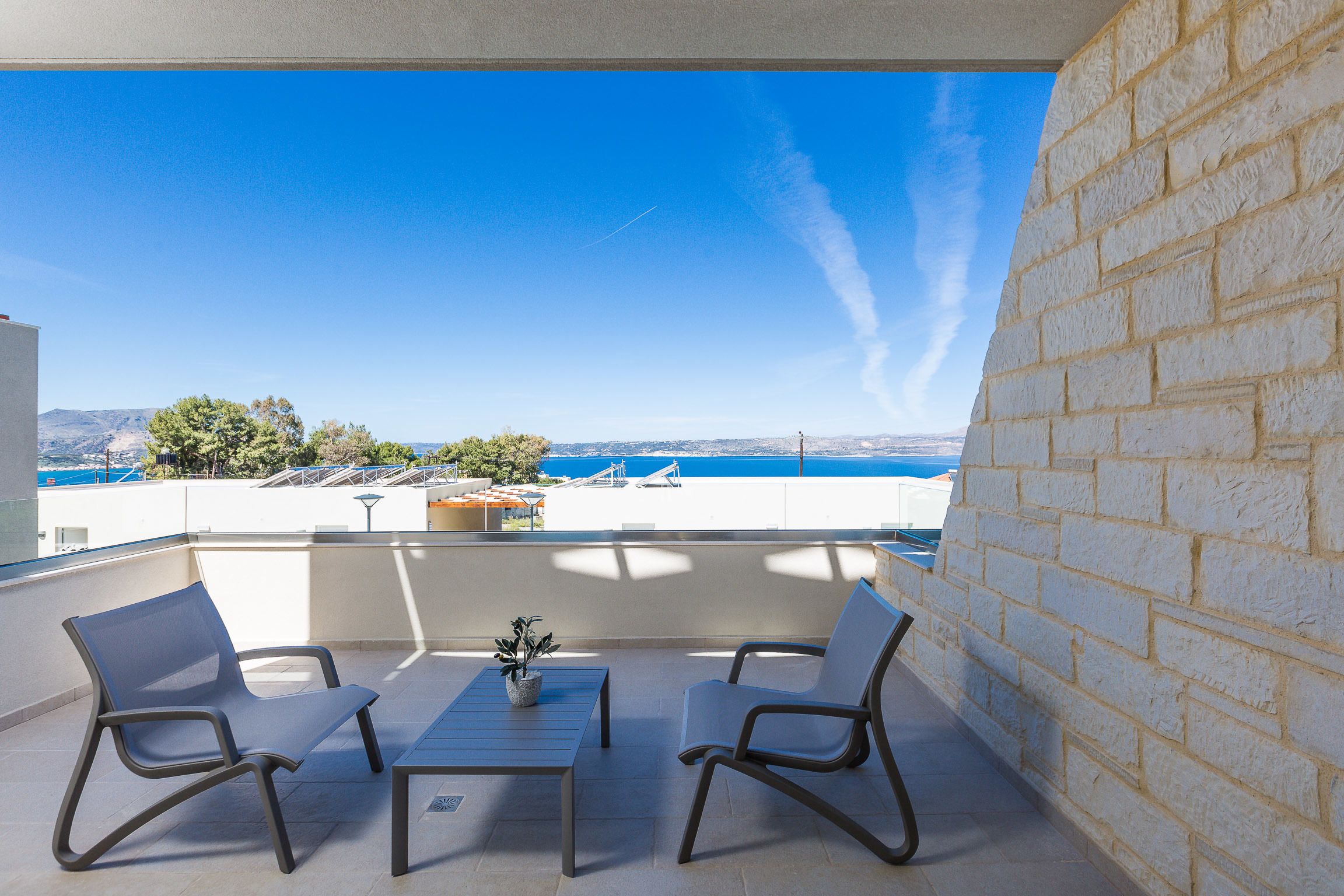 A balcony with two chairs and a small table, overlooking a scenic view of the sea and distant mountains.