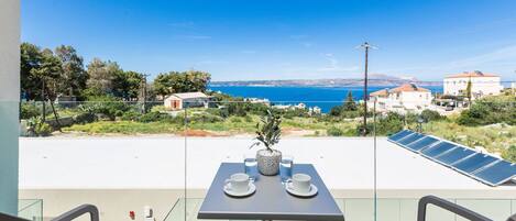 A balcony with a table set for two, overlooking a coastal area with houses and a clear blue sky.