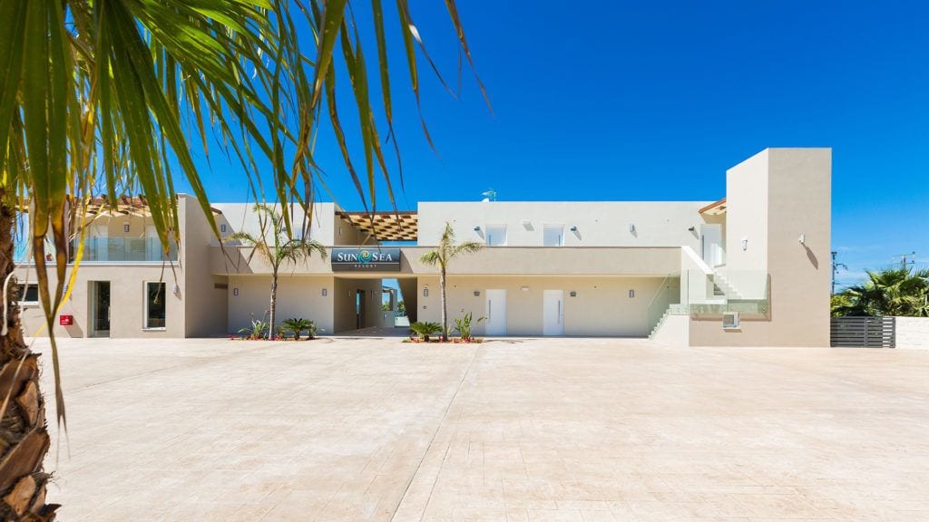 Un hotel moderno con entrada central, palmeras y un cielo azul despejado.