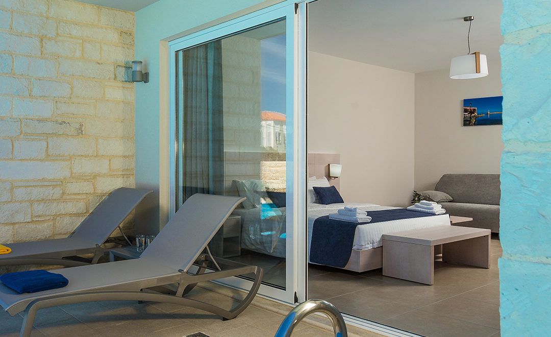 Quarto de hotel moderno com porta de vidro que dá para uma varanda, cama com colcha azul, sofá cinza e uma mesa pequena.