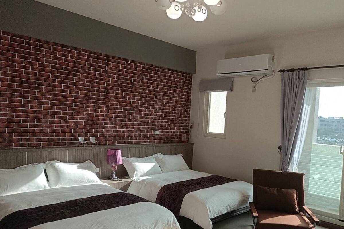 Une chambre d’hôtel avec deux lits, un mur en briques, un appareil de climatisation, une fenêtre avec des rideaux et un balcon.