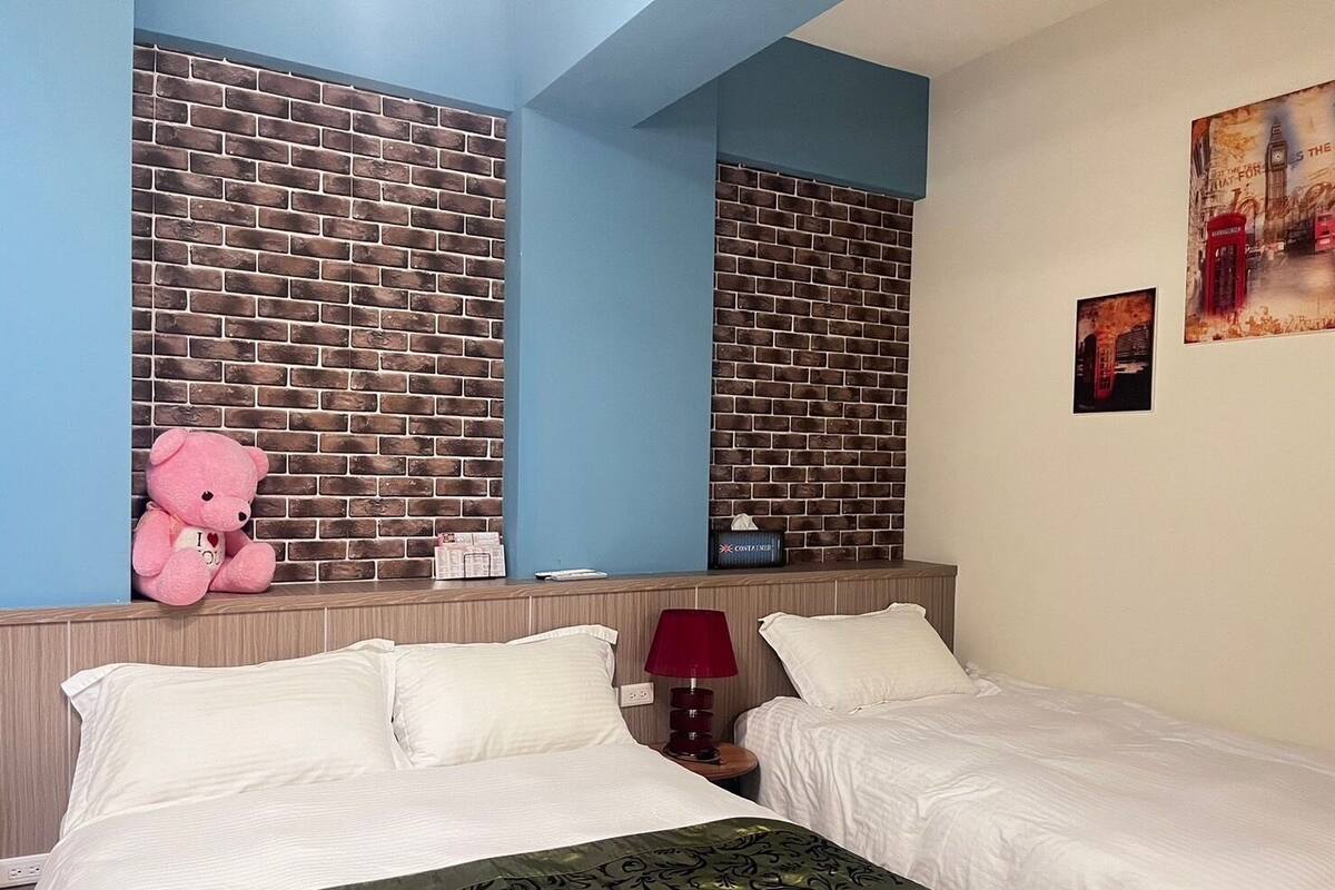 Une chambre d’hôtel avec deux lits, un mur décoré d’un motif en briques, un ours en peluche, une lampe rouge et un tableau représentant une cabine téléphonique rouge.