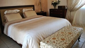 A neatly made bed with a floral-patterned bench, a bedside table with a lamp, and a dresser with a vase of flowers. - Paradise Villa Sounio (Saronikos)